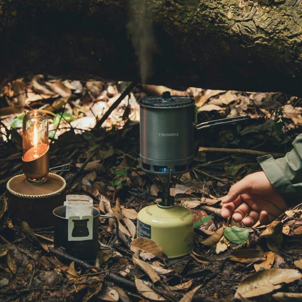 Camping scene with a coffee maker, lantern, and person's hand in a forest setting.