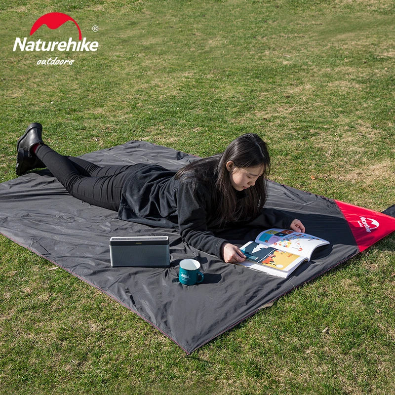 Person lying on a large gray outdoor blanket reading a book with a Naturehike logo in the corner.