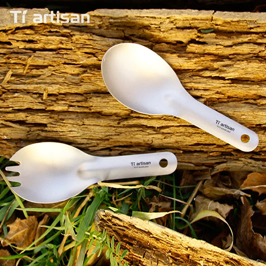Two titanium spoons with 'Ti artisan' branding on a wooden surface with grass and leaves.