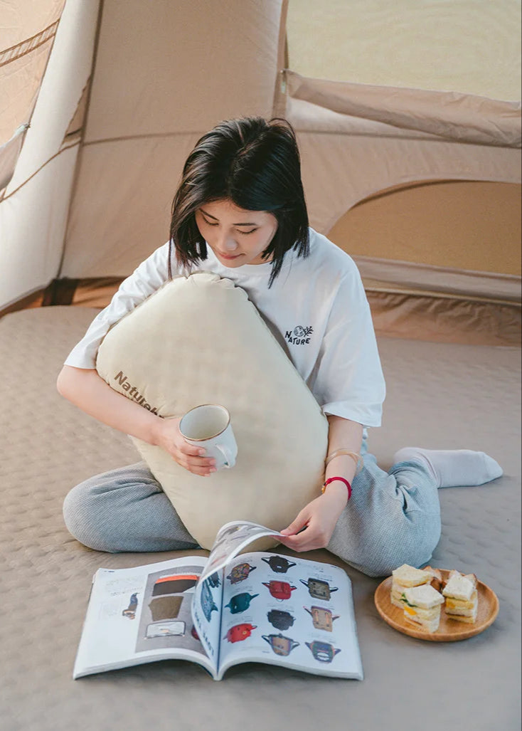 Person sitting on the floor with a large cushion, reading a book and holding a cup.