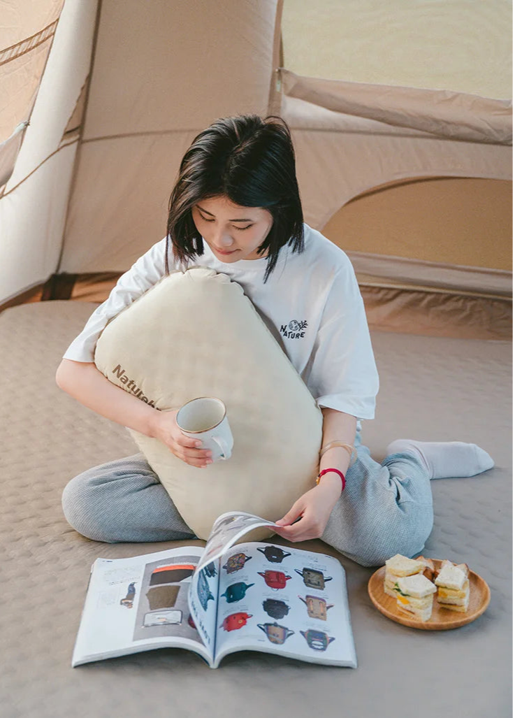 Person sitting on the floor with a large cushion, reading a book and holding a cup.