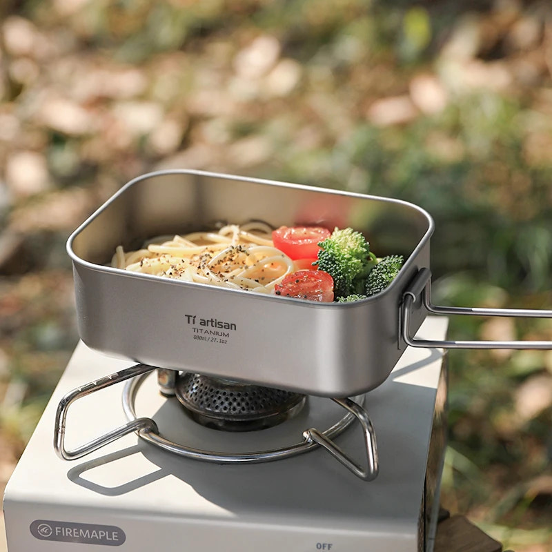 Portable stove with a pot containing noodles, tomatoes, and broccoli outdoors.
