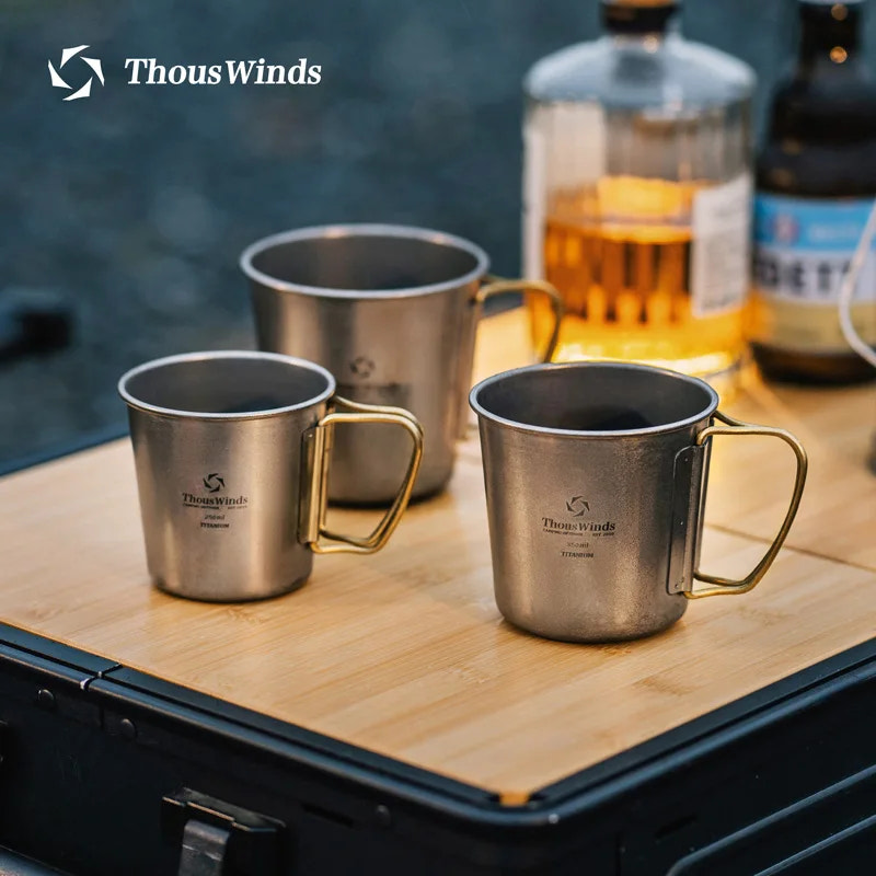 Three metal mugs with gold handles on a wooden surface, featuring the ThousWinds brand.