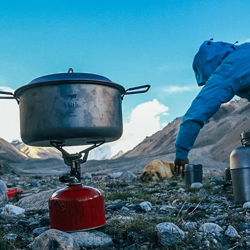 Person in blue jacket using a camping stove with a pot outdoors in a mountainous area.