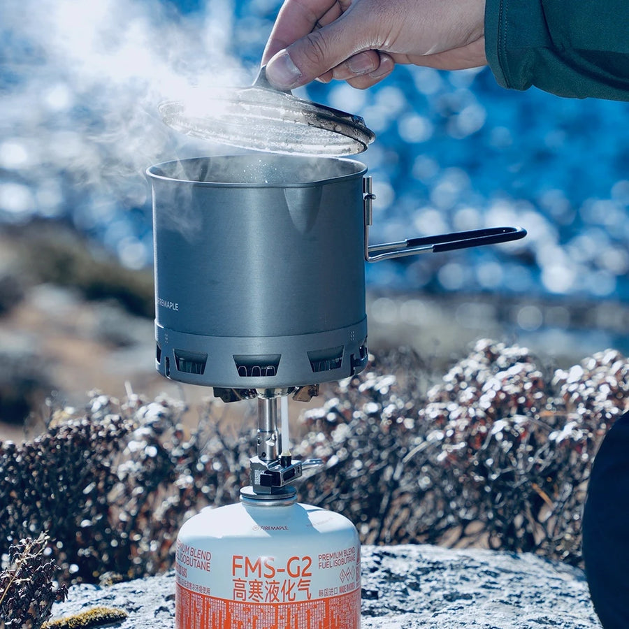 Person using a portable stove with a pot outdoors, surrounded by snow and rocks.