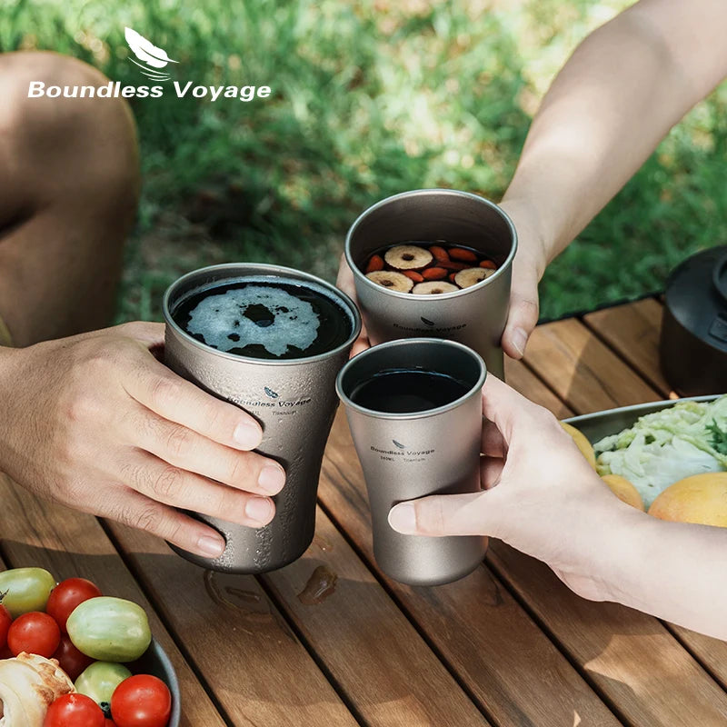 Three people holding Boundless Voyage travel mugs outdoors on a wooden table.