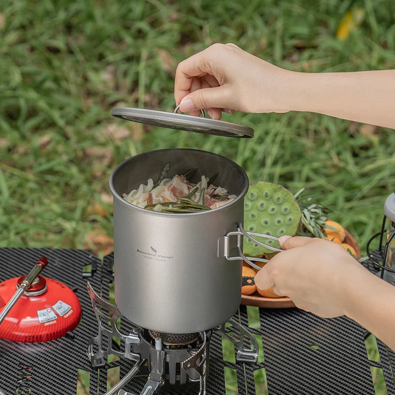 Person cooking outdoors using a portable stove and pot with food on a grassy background