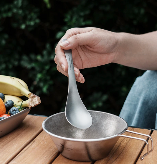 Person holding a titanium spoon over a metal bowl with a blurred outdoor background