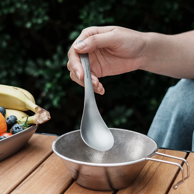 Person holding a titanium spoon over a metal bowl with a blurred outdoor background