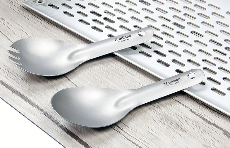 Two titanium spoons with 'Ti artisan' branding on a wooden surface.