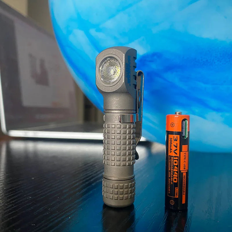 Small flashlight next to a battery on a reflective surface with a blue background