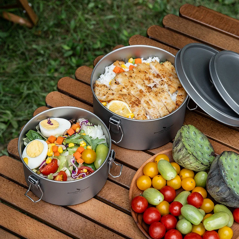 Two metal food containers with meals on a wooden table outdoors.