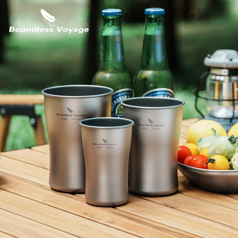 Three titanium cups on a wooden table with Boundless Voyage branding, surrounded by bottles and fruits.