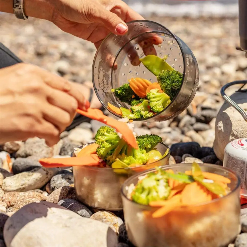 Person preparing vegetables on a rocky beach with a portable stove.